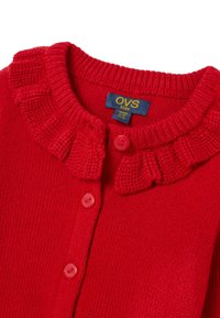 Rode gebreide cardigan met een gerimpelde kraag, vier roze knopen en een blauw label met gele tekst die de maat aangeeft.