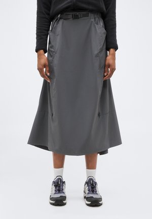 Άτομο που φοράει μεσαίο φόρεμα ανθρακί γκρι με πλαϊνές τσέπες με φερμουάρ, μαύρο μακρυμάνικο τοπ, λευκές κάλτσες και γκρι αθλητικά παπούτσια.