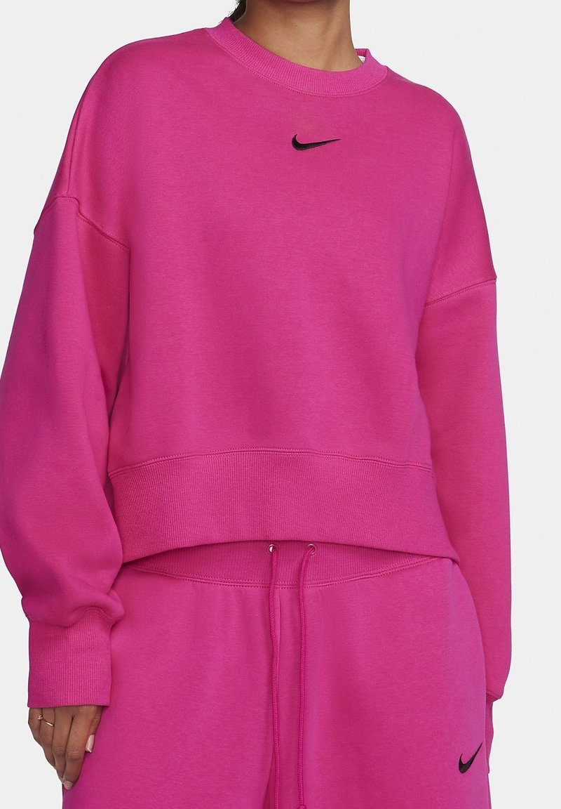 Personne portant un sweatshirt Nike rose vif et un pantalon de survêtement assorti, les deux comportant de petits logos Swoosh Nike noirs.