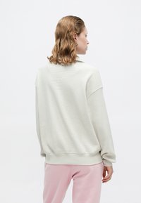 Sweatshirt tricotat, texturat, de culoare gri deschis, cu umeri căzuți și mânici cu elastic, asortat cu pantaloni de trening roz, prezentat din spate.