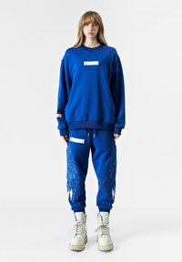 Sudadera azul con un panel de acento blanco, combinada con pantalones jogger azules que tienen un intrincado bordado de alas y acentos blancos. Botas blancas completan el conjunto.