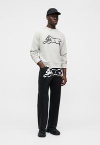 Ανοιχτό γκρι φούτερ με μαύρο αφηρημένο σχέδιο γάτας, σε συνδυασμό με μαύρο παντελόνι που έχει παρόμοιο λευκό περίγραμμα γάτας και μαύρα αθλητικά παπούτσια.