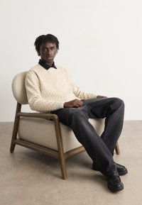 Pulover cu model crem și guler negru, așezat într-un fotoliu cu cadru din lemn și tapițerie bej, purtând blugi negri largi și încălțări.