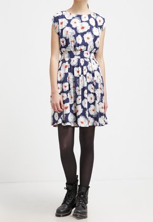 Frau trägt ein marineblaues Kleid mit weißem und orangefarbenem Blumenmuster, schwarze Strumpfhosen und schwarze Schnürstiefel, steht vor weißem Hintergrund.