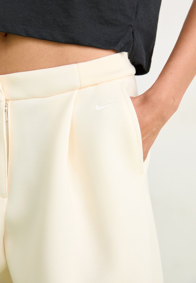 Persona in pantaloni Nike off-white con una mano in tasca e una maglietta nera corta, che mostra il ventre e i dettagli della cintura.