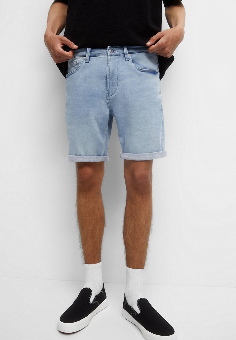 Shorts en denim bleu clair avec des revers, présentant un design classique à cinq poches, portés avec des chaussures noires à enfiler et des chaussettes blanches.