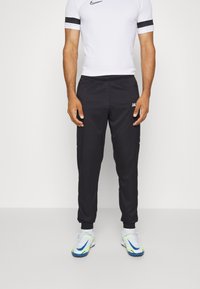 Zwarte sportbroek met een elastische tailleband, voorzien van een wit logo en smalle boorden. Gdragen met een witte fitted shirt en sportschoenen.