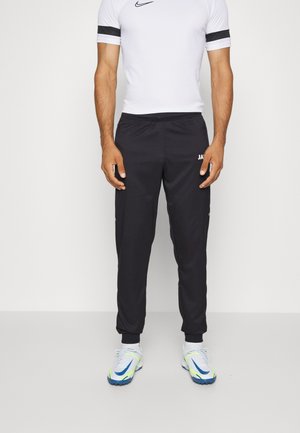 Pantalons de sport noirs avec une taille élastique, dotés de détails en logo blanc et de poignets fuselés. Portés avec une chemise blanche ajustée et des chaussures de sport.