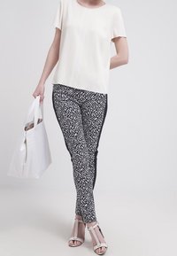 Vit, kortärmad topp i kombination med leggings med svart och vitt mönster. Modellen håller en vit tygväska och har på sig vita sandaler med remmar.