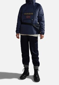 Marinblå pufferjacka med en ficka med dragkedja, logotyp-patch och huva; kombinerad med mörka cargobyxor och svarta snörstövlar med tjocka sulor.