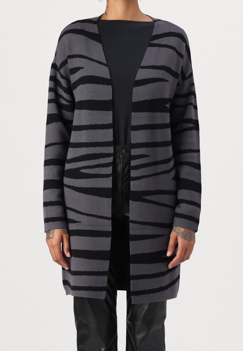 Szary i czarny kardigan w paski, otwarty z przodu, z długimi rękawami, o miękkiej fakturze, do połowy długości, noszony na czarnym topie i czarnych spodniach.