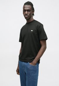 Juoda trumpomis rankovėmis medvilninė marškinėliai, ant krūtinės su mažomis žaliomis Lacoste logotipu, derinami su mėlynais džinsais.