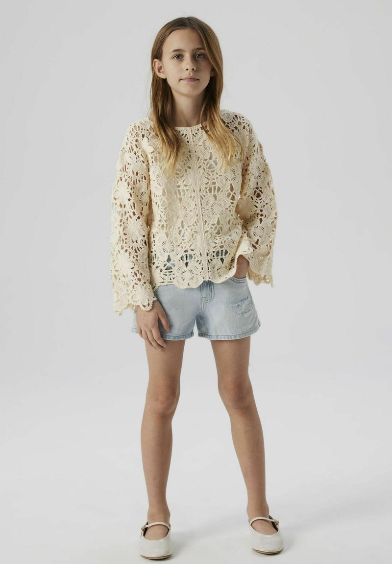 Top in crema all'uncinetto con maniche lunghe, caratterizzato da motivi floreali e orlo scallop, abbinato a pantaloni corti in denim chiaro e scarpe bianche.