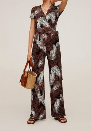 Femme portant une combinaison marron et blanche à motif de feuilles, tenant un sac à main en paille tressée avec des poignées en cuir, et portant des sandales marron.