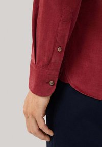 Rode corduroy shirtmanchet met twee ronde knopen, die de gestructureerde stof en de elegante, getailleerde pasvorm toont. Hand zichtbaar tegen donkere stof.