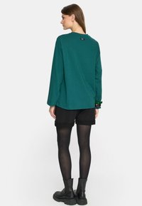 Zielona bluzka z długim rękawem, wykonana z miękkiego materiału, czarne szorty, czarne rajstopy i masywne czarne botki. Posiada okrągły dekolt oraz detal z logo z tyłu.