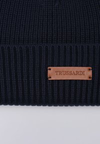 Cappellino a maglia blu navy con texture a coste; presenta un'etichetta in pelle marrone con la scritta "TRUSSARDI" in rilievo.