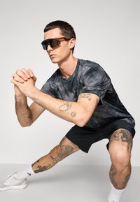 Mand i sort træningsoutfit og solbriller, med tatoveringer på arme og ben, udfører en sidelæns strækning mod en enkel baggrund.