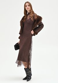 Bruine kanten jurk met een doorschijnende textuur, lange mouwen en een gelaagde zoom. Gecombineerd met een donkerbruine jas gevoerd met nep bont en zwarte enkellaarsjes met hak.