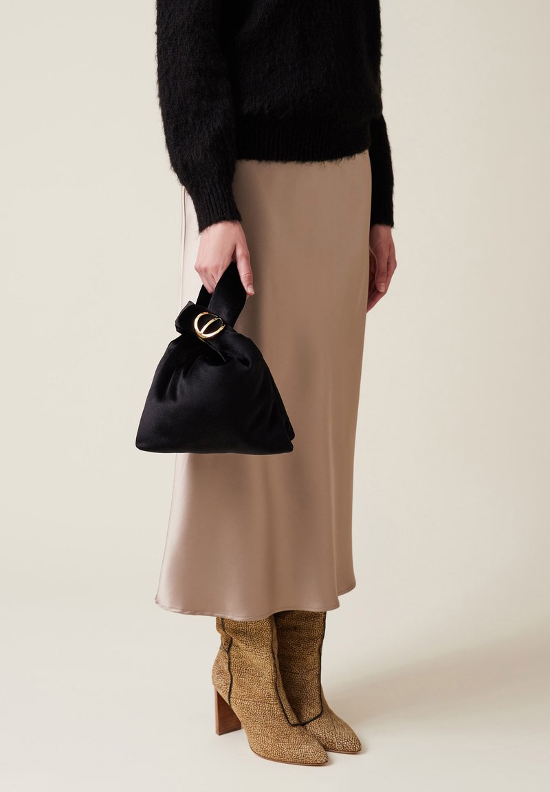 Borsa nera in raso con parte superiore annodata e dettagli in oro. Una gonna in raso beige aderente e stivali alla caviglia beige con motivo completano il look.