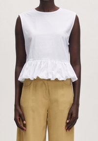 Blouse blanche sans manches avec un ourlet peplum froncé portée avec un pantalon taille haute beige sur une personne à la peau foncée devant un fond clair.