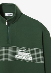 Sweat-shirt vert avec un col montant, une fermeture éclair, et un motif de grille blanche comportant un logo crocodile brodé ainsi que le texte "Le Club Lacoste".