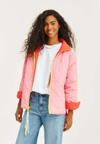 Prošívaná růžová bunda s oranžovými akcenty, žlutým zipem a strukturální povrchovou úpravou. V kombinaci s bílým tričkem a modrými džíny.