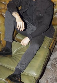 Zwarte spijkerjack over een zwart shirt, grijze slim-fit broek en zwarte schoenen met wingtip-accenten, zittend op een groene leren bank.