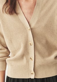 Cardigan en maille beige texturée avec quatre boutons fixés, porté sur une jupe plissée noire, montrant le milieu du torse et les manches retroussées.