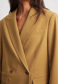 Mujer vistiendo una chaqueta color mostaza de doble botonadura con solapas muestradas y bolsillos de solapa, visible desde el cuello hasta la cintura.