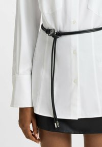 Black rope belt with silver hardware, worn over a white button-up shirt. Belt features a knot design, ends finished with metal tips.