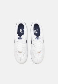 Bijele Nike Air Force 1 tenisice s mornarsko plavim akcentom, koje imaju perforiranu prednju stranu, okrugle vezice i prepoznatljiv logo na jeziku.