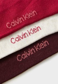 Tre tygprover i röd, krämfärgad och mörkröd, med texten "Calvin Klein" vävd i en kontrasterande nyans på varje.