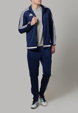 Marineblauw sporttrainingspak met witte strepen op de mouwen en broek, voorzien van een doorritsjack en lichte stof. Grijze T-shirt eronder.