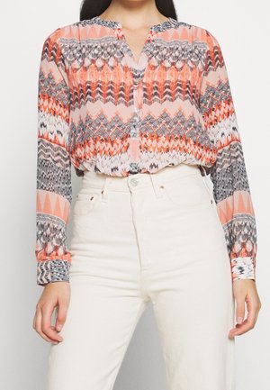 Femme portant un chemisier à manches longues à motifs géométriques orange, noir et blanc, rentré dans un pantalon blanc taille haute.