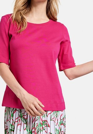 Fuchsia Baumwoll-T-Shirt mit Rundhalsausschnitt und kurzen Ärmeln, versehen mit einem dezenten Logo am Saum. Kombiniert mit einem gemusterten Rock.