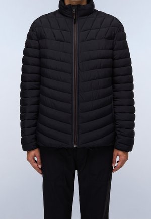 Mann trägt eine schwarze gesteppte Jacke mit durchgehendem Reißverschluss, horizontalen und Chevron-Nähten, sowie schwarze Hosen vor hellem, einfarbigem Hintergrund.