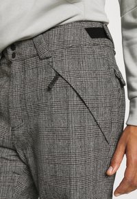 Pantalon à carreaux gris avec un tissu texturé, une poche latérale inclinée et un panneau élastique noir contrastant à la ceinture.