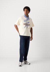 Beige kortærmet skjorte med blomster grafik og "HEATHE" tekst, parret med mørkeblå loose-fit jeans og et blåt bandana. Sølvfarvede sneakers fuldender looket.