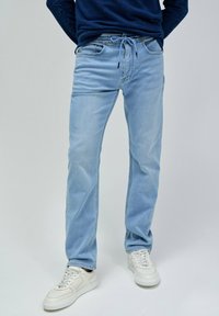 Jeans de mezclilla azul claro con un ajuste Slim, cintura elástica, cierre de cordón y bolsillos frontales. Combinados con zapatillas blancas.