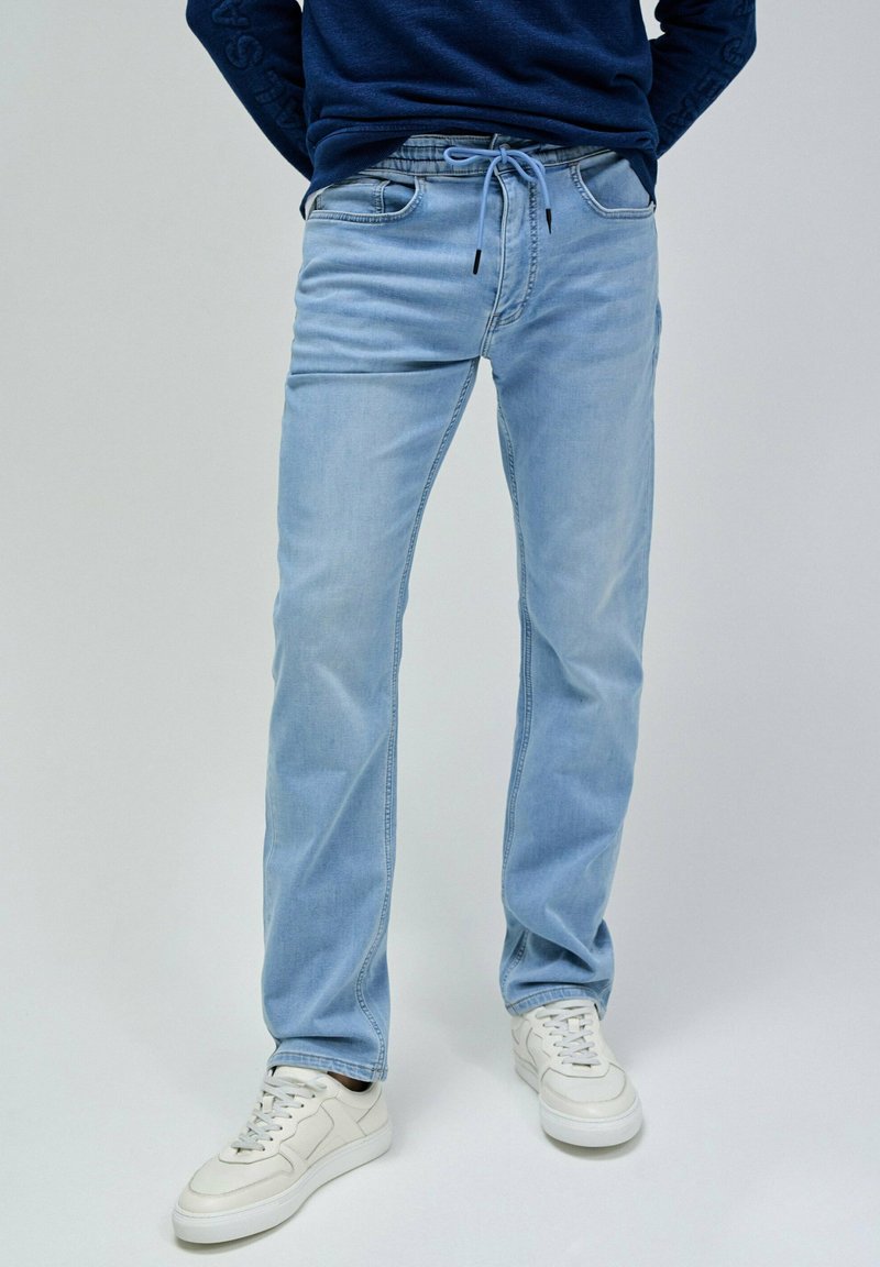 Jeans de mezclilla azul claro con un ajuste Slim, cintura elástica, cierre de cordón y bolsillos frontales. Combinados con zapatillas blancas.