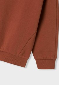 Bruine sweatshirt detail met ribbels aan de manchetten, accentuerende zijsplitten en een zachte textuur. Focus op materialen en stiknaden.