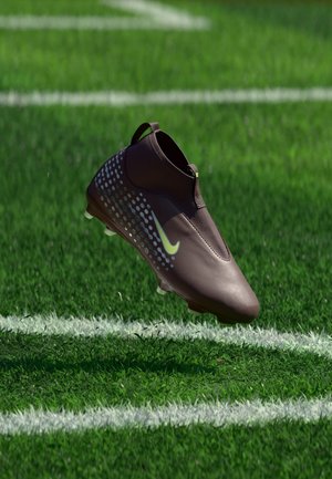 Chaussure de football marron foncé avec des points bleus et un logo Nike jaune, flottant au-dessus de l’herbe verte près des lignes blanches du terrain.