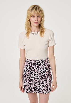 Lichtbeige nauwsluitend topje met korte mouwen, gecombineerd met een roze rok met een zwart gespikkeld patroon. Accent van een zilverkleurige ronde ketting.