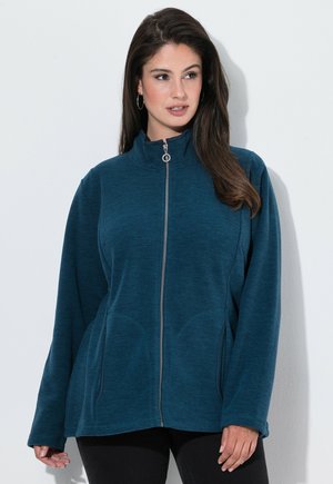 Mujer con chaqueta polar azul con cremallera y bolsillos laterales, de pie sobre un fondo blanco, mirando hacia su izquierda.