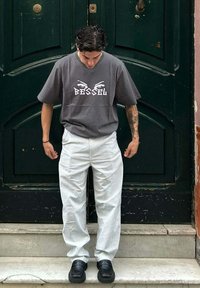 Camiseta de algodón gris con gráfico "BESSEL" y diseño de ojos; pantalones a rayas blancas, zapatos gruesos negros, de pie en unos escalones frente a una puerta verde.
