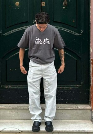 Camiseta de algodón gris con gráfico "BESSEL" y diseño de ojos; pantalones a rayas blancas, zapatos gruesos negros, de pie en unos escalones frente a una puerta verde.