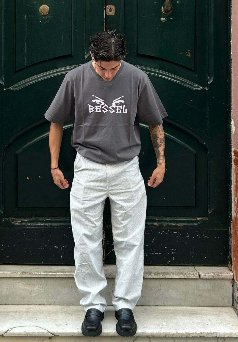Camiseta de algodón gris con gráfico "BESSEL" y diseño de ojos; pantalones a rayas blancas, zapatos gruesos negros, de pie en unos escalones frente a una puerta verde.