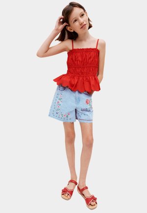 Niña con top sin mangas rojo con volantes, pantalones cortos de mezclilla azul claro bordados y sandalias rojas, de pie con la mano tocándose el cabello.