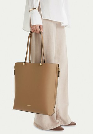 Žena držící velkou hnědou koženou tašku, oblečená ve světlých béžových širokých kalhotách, bílé blůze, hnědých špičatých botách a zlatém náramku.